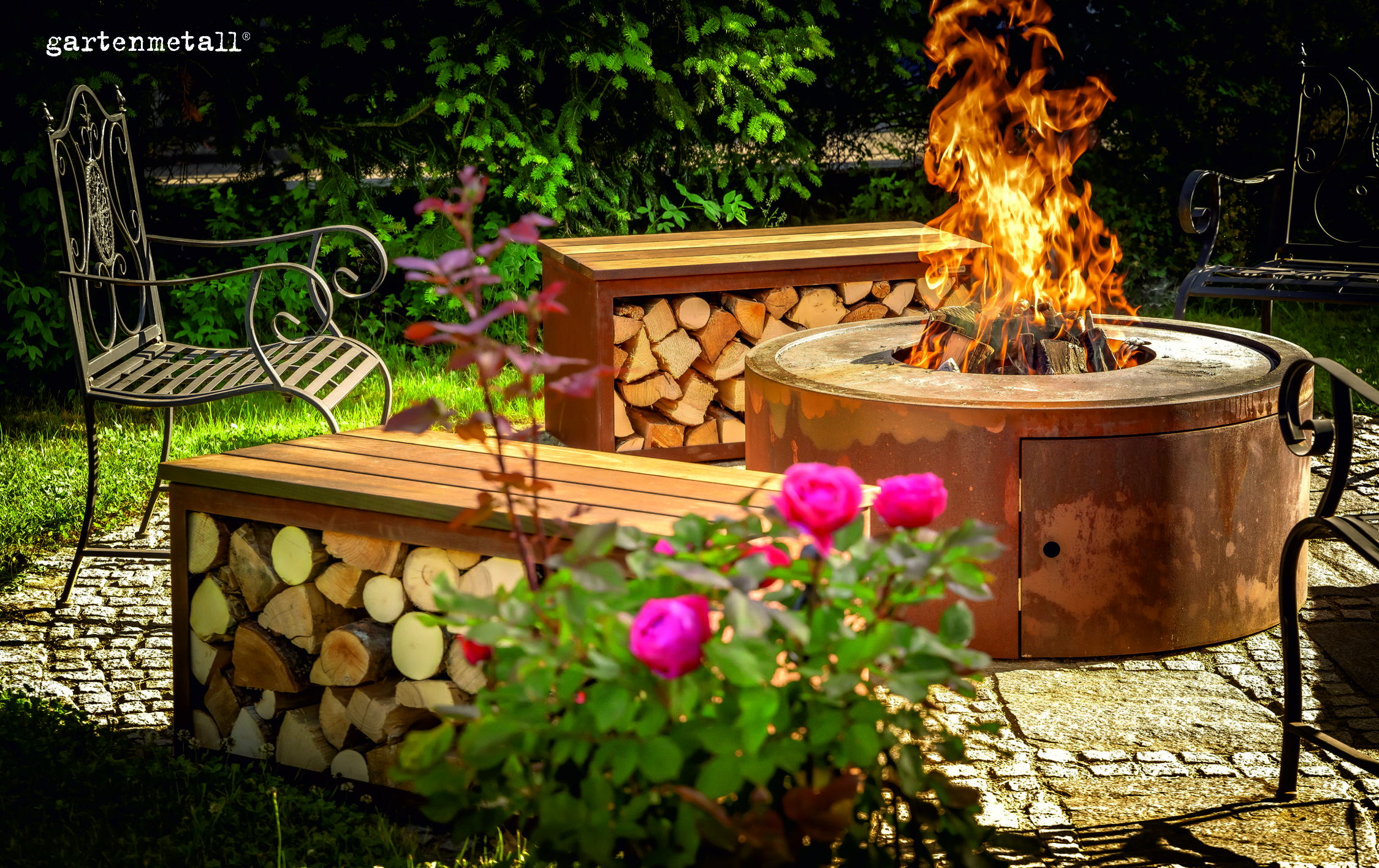 Moderne Runde Feuerstelle aus Cortenstahl mit brennenden Holzscheiten und zwei Bänken aus Cortenstahl mit Holzsitzflächen.