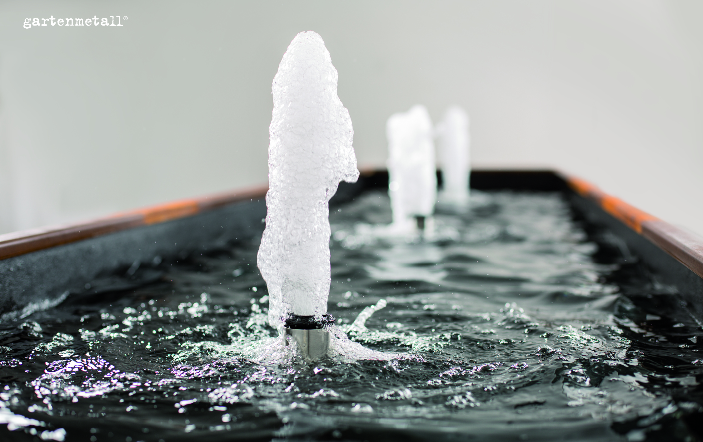 Wasserfontäne sprudelt lebhaft in rechteckigem Becken, zwei weitere Fontänen im Hintergrund. Oberflächenwasser reflektiert Licht, ruhiges, dezentes Ambiente. Text: 'gartenmetall®'.