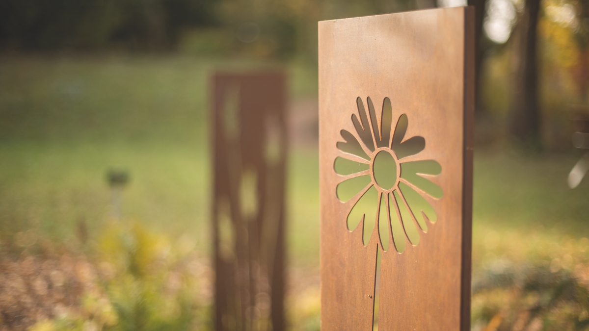 Metallplatte zeigt stilisierte Blume, stehend im Sonnenlicht. Im Hintergrund unscharfe Naturkulisse mit Wiese und Bäumen. Rustikale Atmosphäre, herbstlicher Frieden vermittelt. Kein Text vorhanden.
