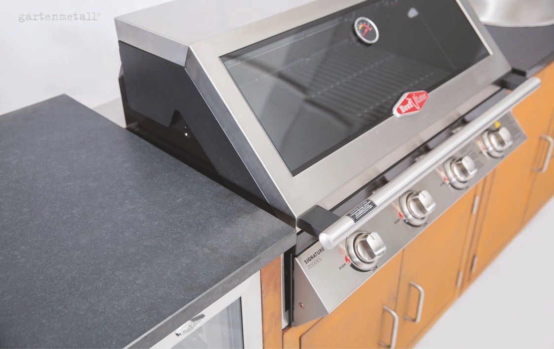 Grill: Ein moderner Edelstahlgrill mit vier Drehknöpfen steht auf einer Holzkonsole. Text: 'gartenmetall®', 'Signateur 2000E-S', 'BeefEater'.  Kontext: Innenraum mit weißem Hintergrund.