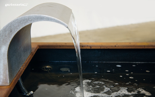 Ein Metallwasserfall fließt in ein rechteckiges Becken, erzeugt Blasen auf der Wasseroberfläche. Links oben steht „gartenmetall®“. Der Hintergrund ist unscharf und schlicht.