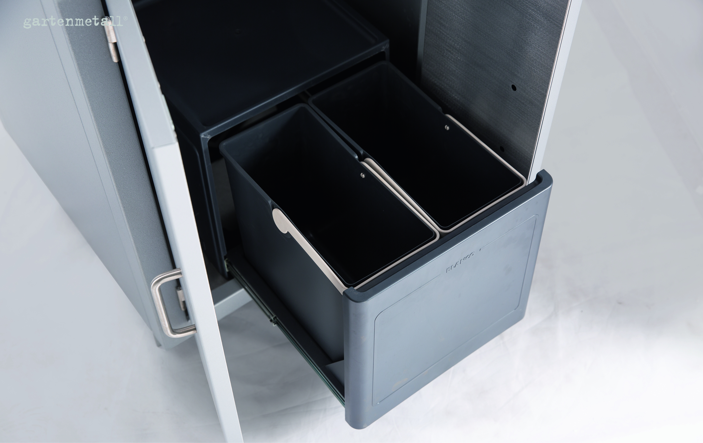 Zwei schwarze Müllbehälter von BLANCO in einem offenen Küchenschrank. Der Schrank ist aus Metall, teilweise geöffnet, um die Behälter herauszuschieben. Oberhalb steht 'gartenmetall'.