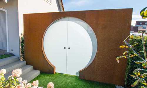 Ein weißes, rundes Tor eingebaut in eine rostige Stahlwand, steht geöffnet in einem Garten mit grüner Wiese und Blumen, neben einem Haus mit einer Treppe.