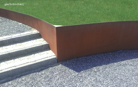 Eine rostfarbene Metallwand trennt eine grüne Rasenfläche von einem steinigen Kiesbereich. Links führen breite graue Steinstufen nach oben. Text: 'gartenmetall'.