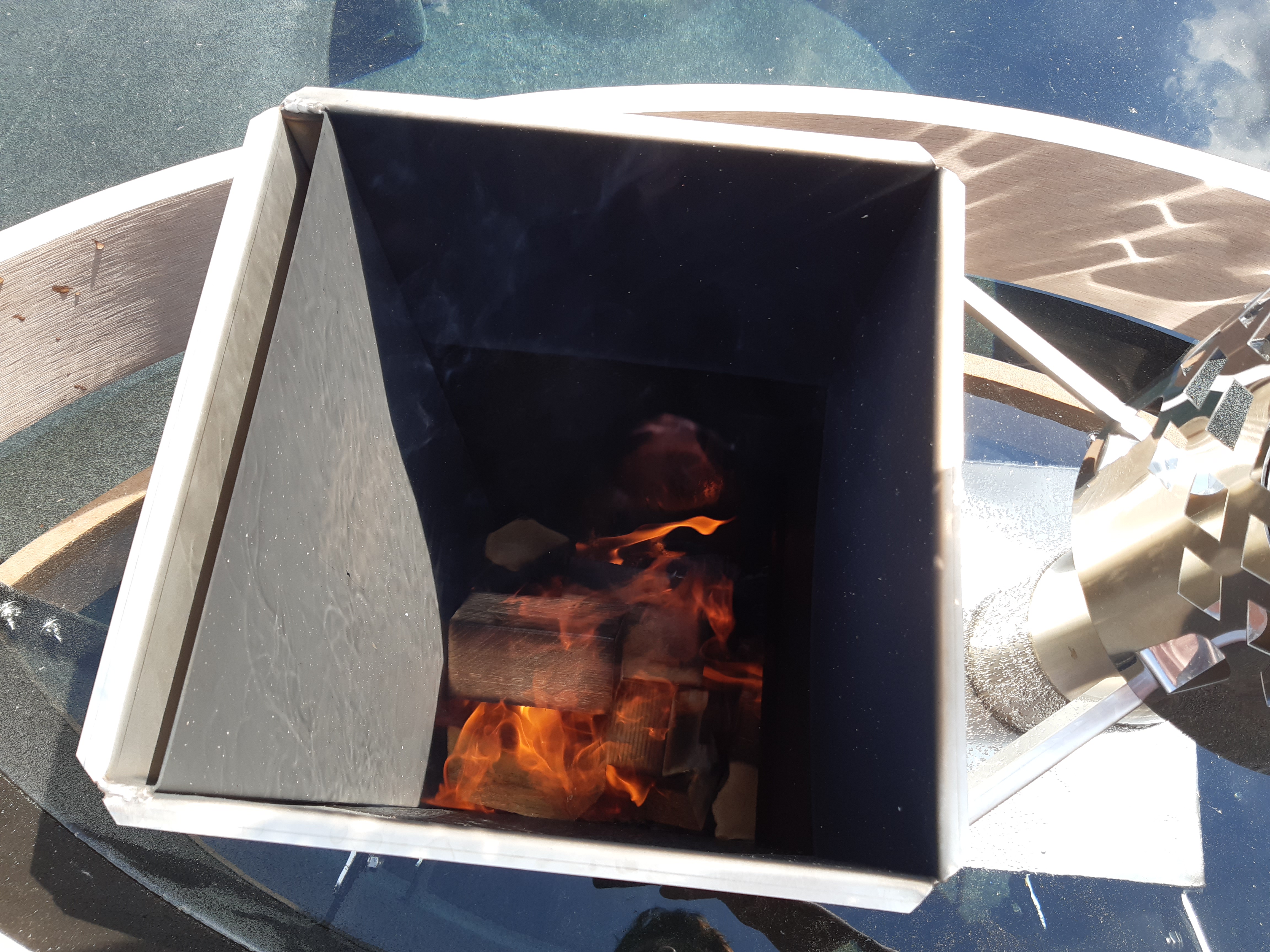 Metalltrichter mit Feuer brennt Holzstücke im Inneren, umgeben von reflektierender Oberfläche bei Tageslicht.