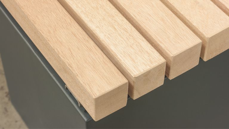 Holzlatten formen eine Sitzfläche auf einer grauen Metallbasis, in einer urbanen Umgebung. Keine sichtbaren Textelemente.