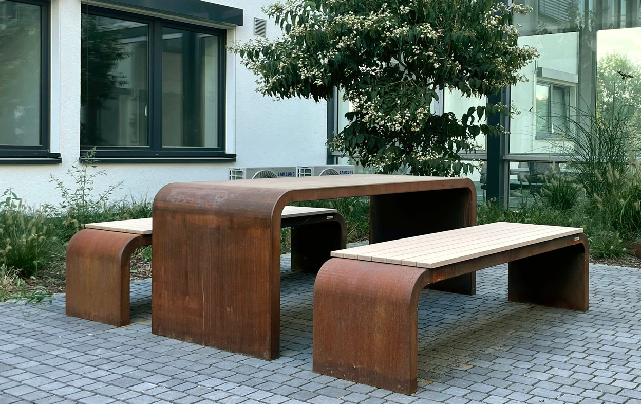 Ein brauner Tisch mit zwei Bänken steht auf gepflastertem Boden, dahinter ein weißes Gebäude mit Fenstern und eine blühende Pflanze in einem Gartenumfeld.