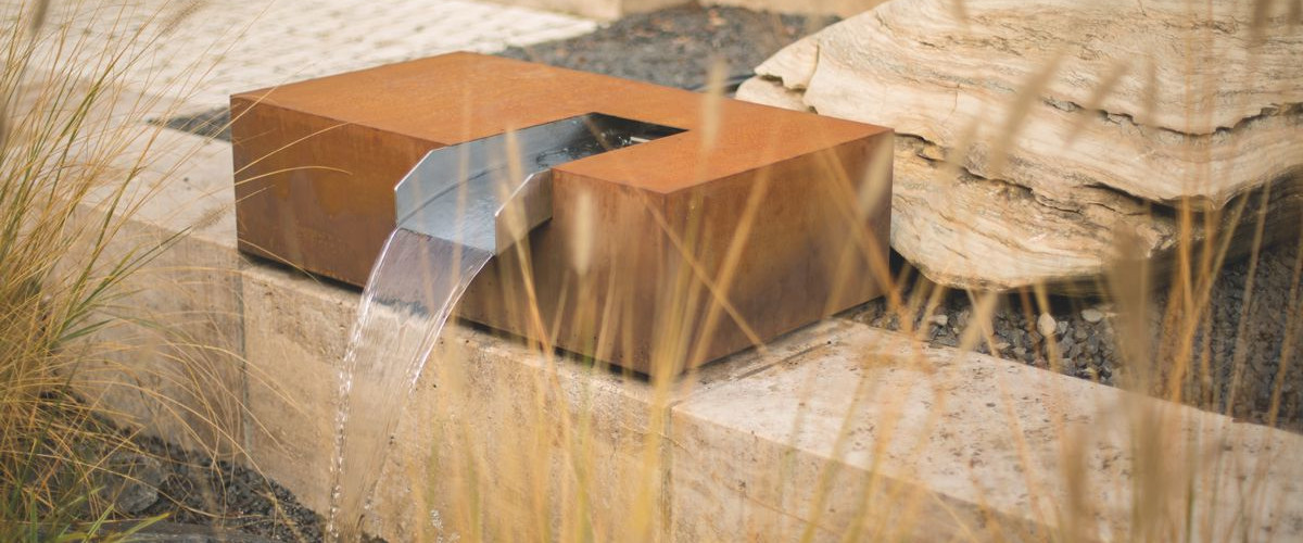 Ein metallischer Wasserspeier fließt stetig in einen kleinen Bachlauf, umgeben von Gras und Steinen, die eine natürliche Landschaftsgestaltung schaffen.