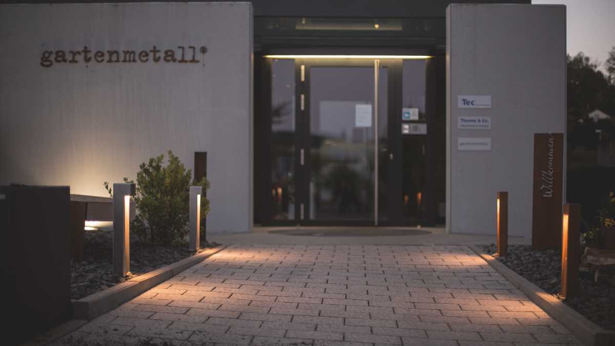 Die Eingangstür eines Gebäudes reflektiert Abendlicht, flankiert von modernen Wegleuchten und begrüntem Umfeld. Schilder: 'gartenmetall®', 'Tec', 'Thomas B. Co.', 'Willkommen'.