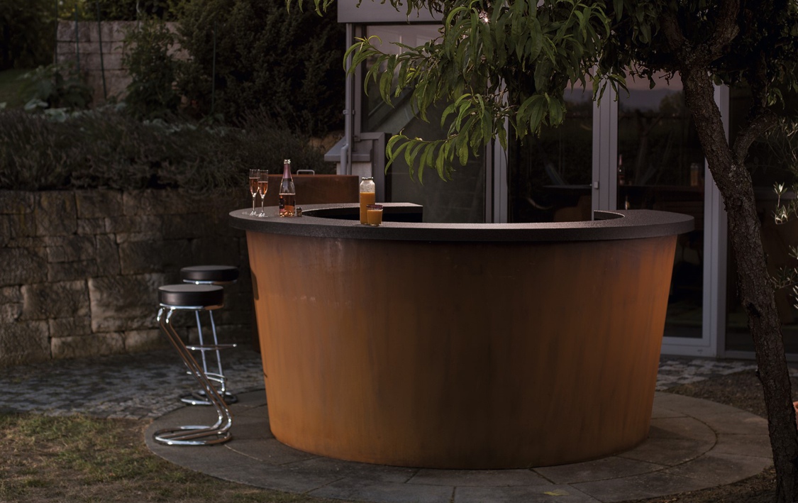 Runde Terrassenbar aus Holz, dekoriert mit Getränken auf der Theke. Im Hintergrund befindet sich eine Mauer und eine moderne Glastür, umgeben von Pflanzen und einem Baum.