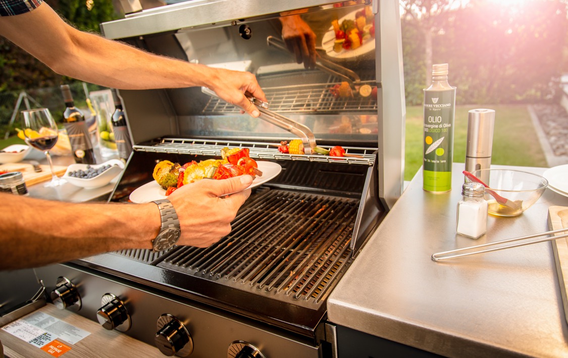 Hände legen mit Zange gegrilltes Gemüse von einem Grill auf einen Teller. Im Garten stehen Olivenöl, Salzstreuer, ein Glas Wein und Beeren auf dem Tisch.