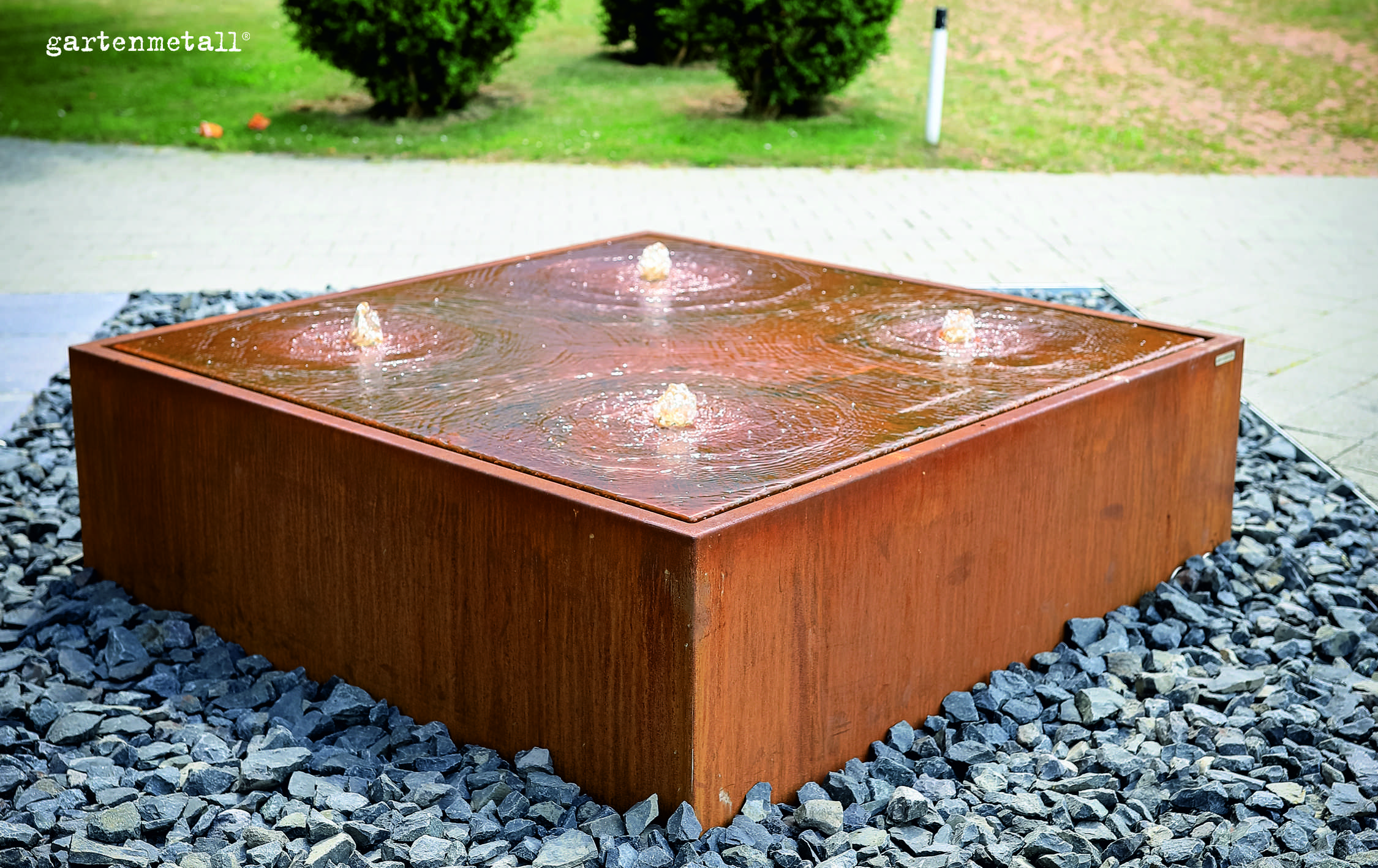 Quadratischer Roststahlbrunnen sprudelt Wasser über vier Düsen, umgeben von grauen Kieselsteinen, auf einem gepflasterten Weg. Text: 'gartenmetall®'.