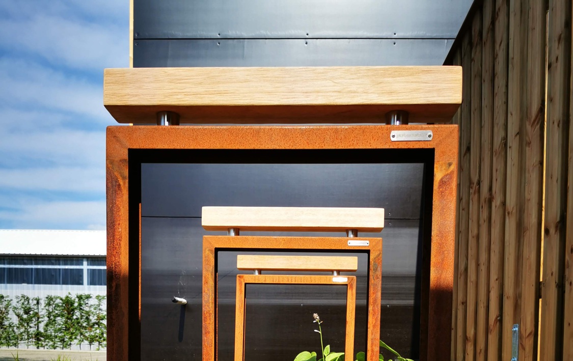 Rostfarbene Metallstrukturen mit Holzleisten wiederholen sich in verschiedenen Größen, vor einer Holzwand im Freien. Dahinter ist ein bewölkter Himmel und ein Gebäudedach erkennbar.