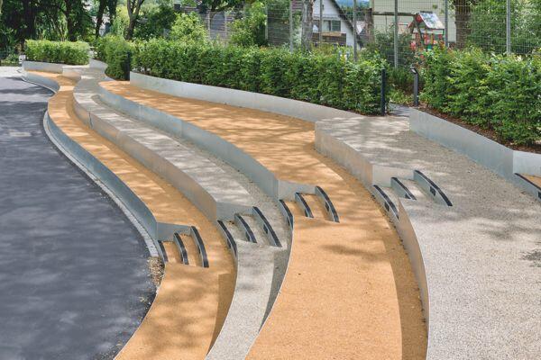 Mehrere geschwungene Sitzstufen aus Beton und Holz ruhen nebeneinander, umgeben von grünen Hecken und Bäumen, neben einem asphaltierten Gehweg in einem Park.