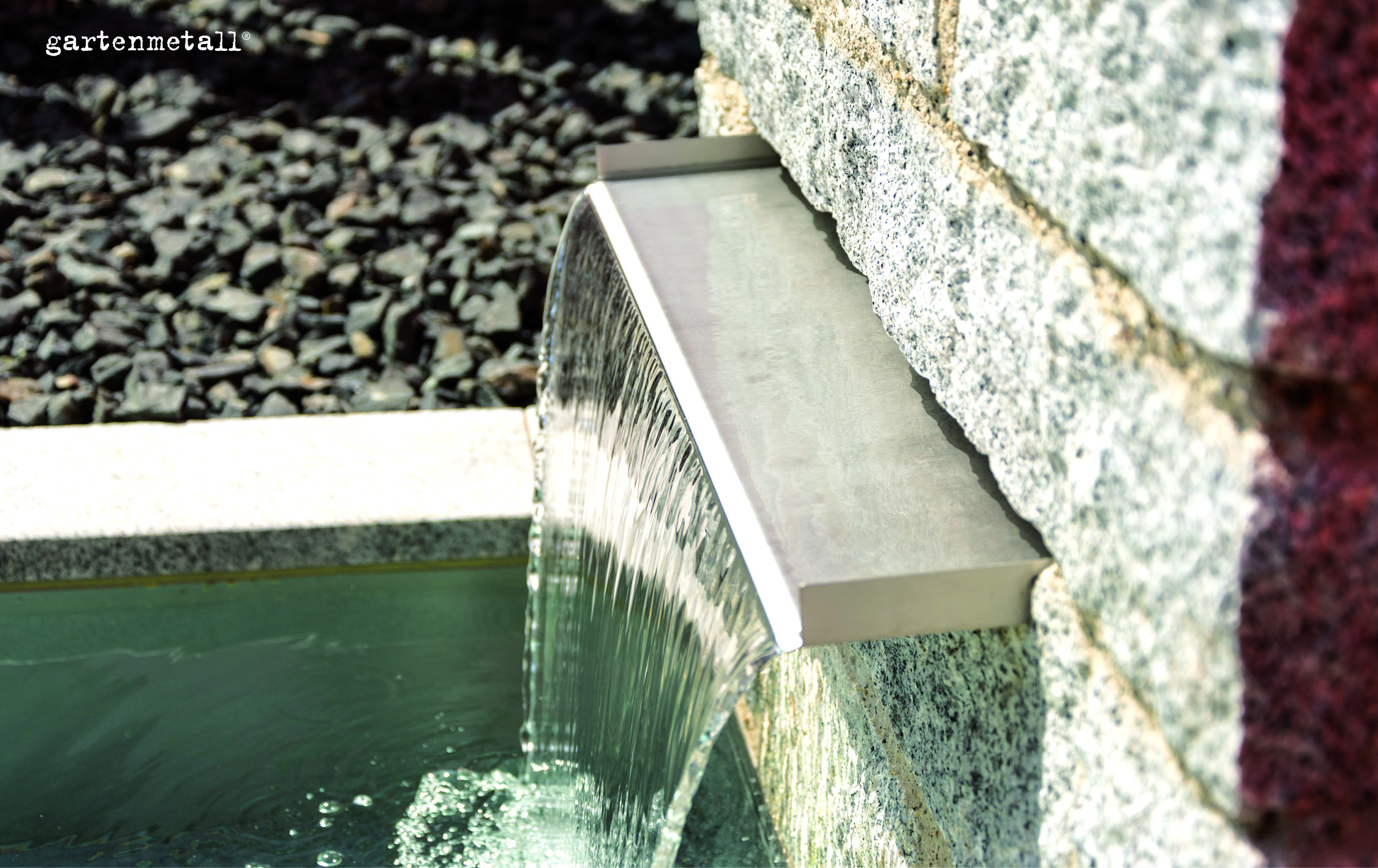 Wasser fließt über einen Metallausguss aus einer Steinmauer in ein Becken, umgeben von dunklen Steinen. Text: 'gartenmetall'.