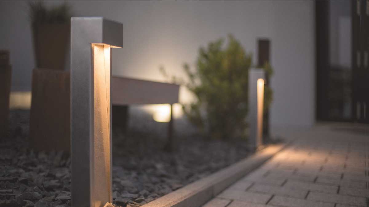 Metallene Sockelleuchten beleuchten einen gepflasterten Weg, flankiert von Kieselsteinen und ein paar Pflanzen vor einem modernen Gebäude.