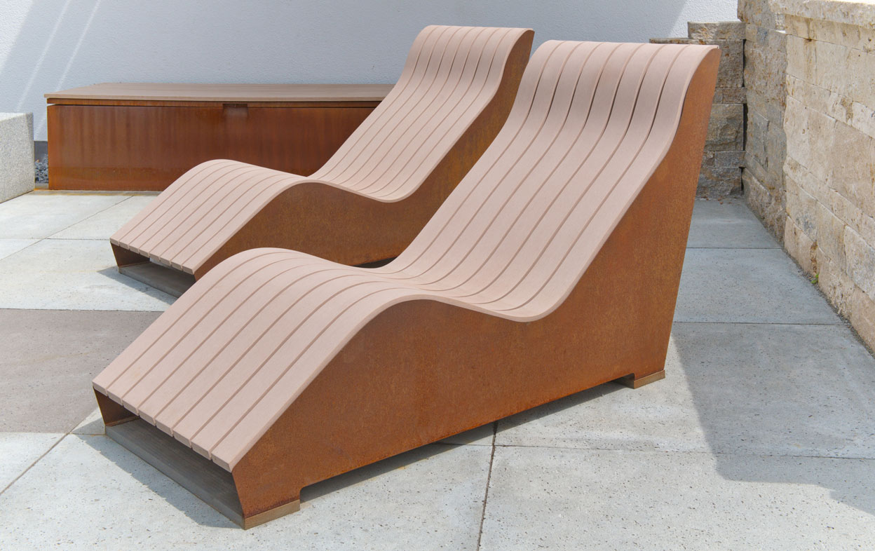 Zwei geschwungene Liegen stehen nebeneinander auf einem gepflasterten Außenbereich. Sie bestehen aus Holzlatten auf rostfarbenen Metallrahmen, umgeben von einer Steinmauer und einer hellen Wand.