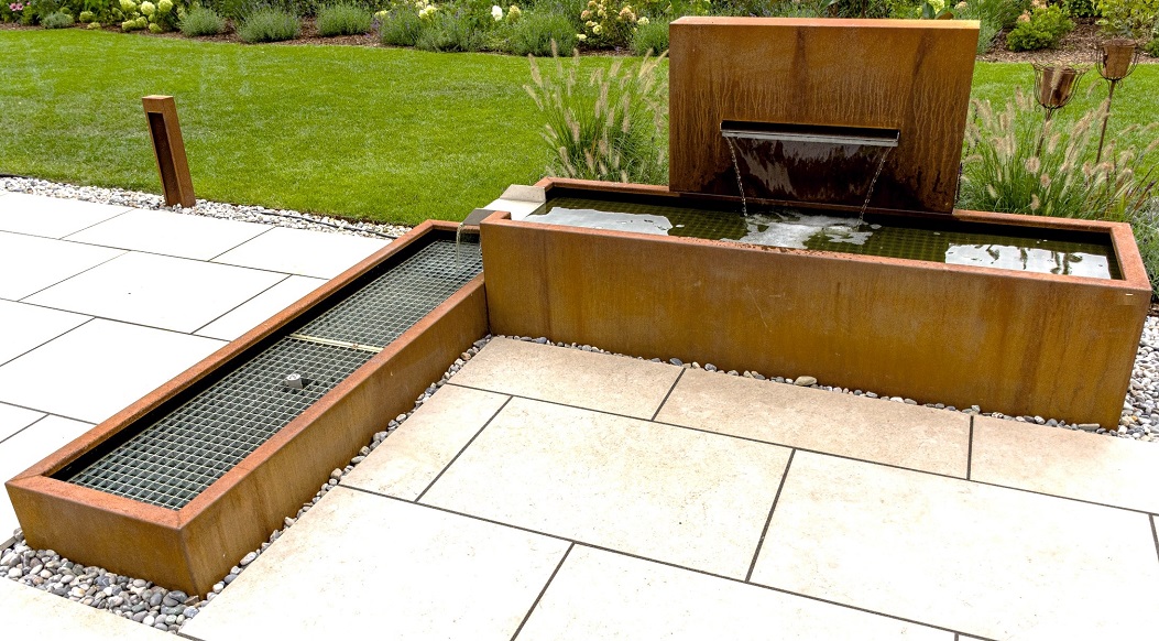 Ein rechteckiger Wasserspeicher aus rostfarbenem Metall fließt in einen kleineren Behälter. Er befindet sich auf einer gefliesten Terrasse, umgeben von gepflegter Rasenfläche und Pflanzen.