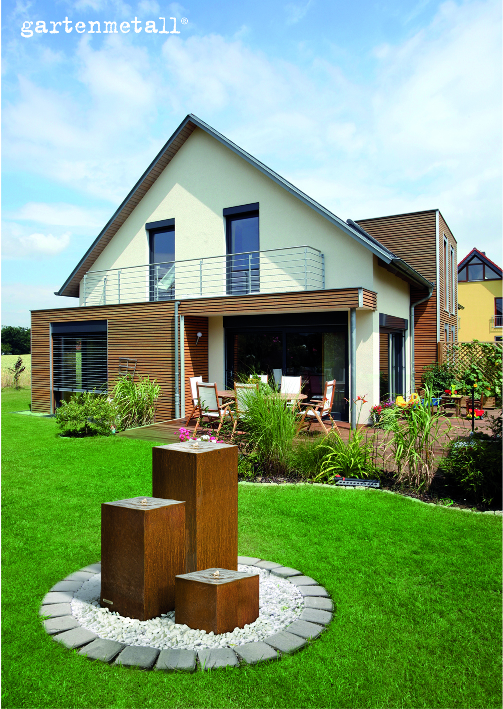 Ein moderner Brunnen mit drei rostfarbenen Säulen sprudelt Wasser in einem Garten, umgeben von grüner Rasenfläche und einem Wohnhaus. Text: 'gartenmetall®'.