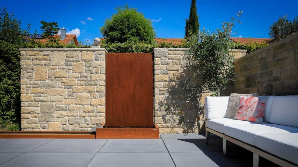 Ein rustikaler Wasserbrunnen fließt an einer Steinwand, umgeben von Pflanzen und modernem Außenmöbel mit weißen Kissen, in einem sonnigen Garten mit blauen Himmel und roten Ziegeldächern im Hintergrund.