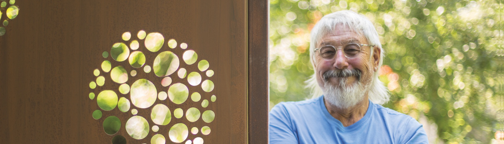 Ein lachender älterer Mann mit weißem Bart steht im Freien neben einer Metallplatte mit runden, durchscheinenden Aussparungen; hinter ihm verschwommene grüne Vegetation.