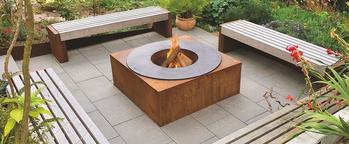 Ein quadratischer Feuerplatz aus rostigem Metall brennt mit Flammen, umgeben von drei Holz-Sitzbänken auf einem gepflasterten Terrassenbereich, mit Pflanzen und Blumen im Hintergrund.