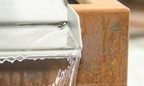 Wasser fließt über die Kante eines rostigen Metallbeckens. Im Hintergrund sind unscharfe Details zu sehen. Text: 'gartenmetall'.