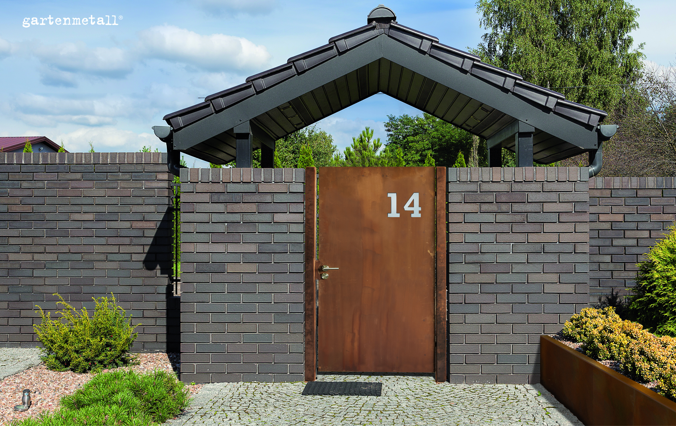 Gate stands still in a brick wall, labeled with the number 14. Surrounded by plants under a blue sky, with the inscription 'gartenmetall®' in the top left corner.