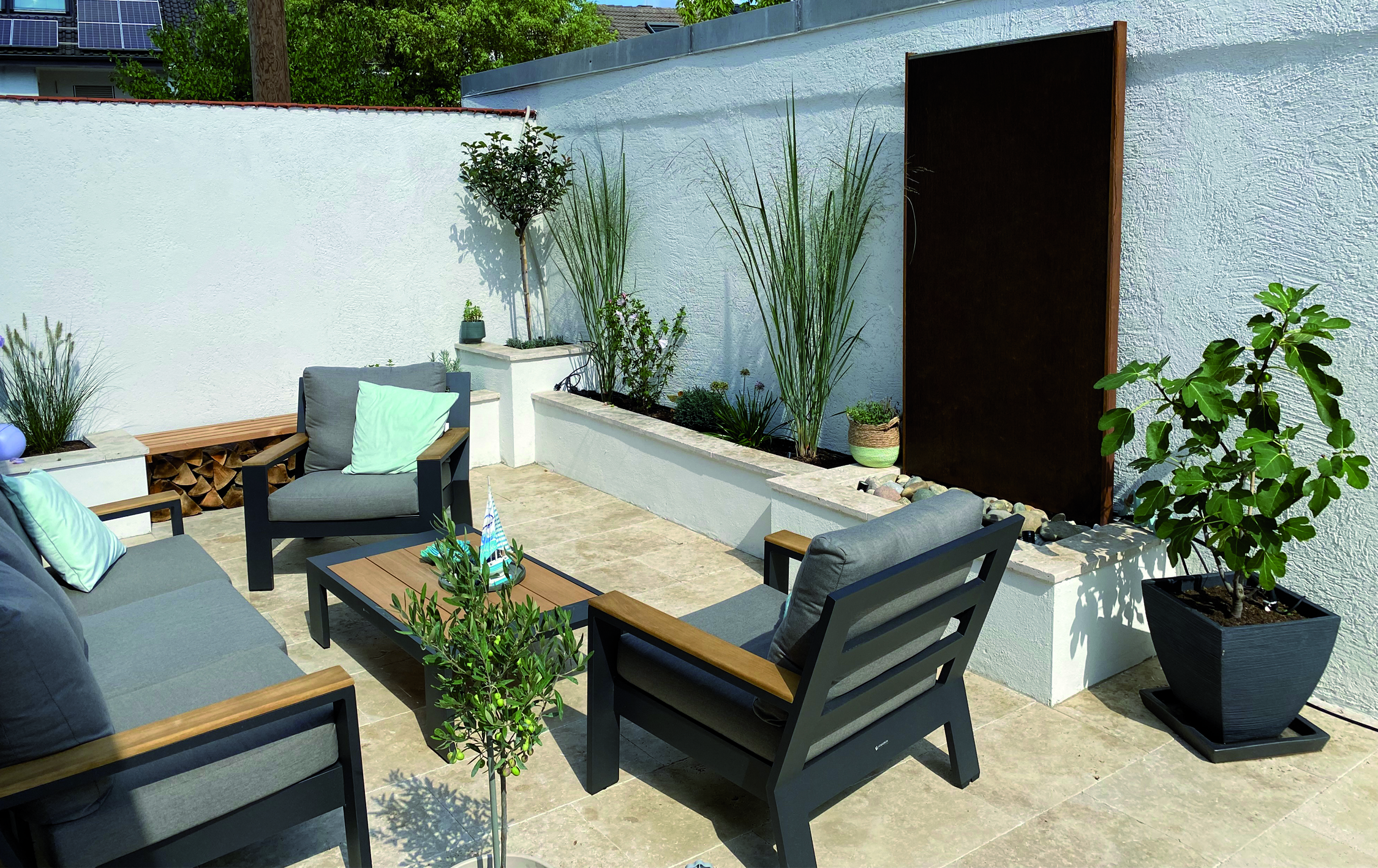 Gartensitzecke mit grauen Sesseln und Tisch, umgeben von Pflanzen und Holzlager an einer weißen Mauer. Ein ruhiger, sonniger Patio mit modernem Ambiente.