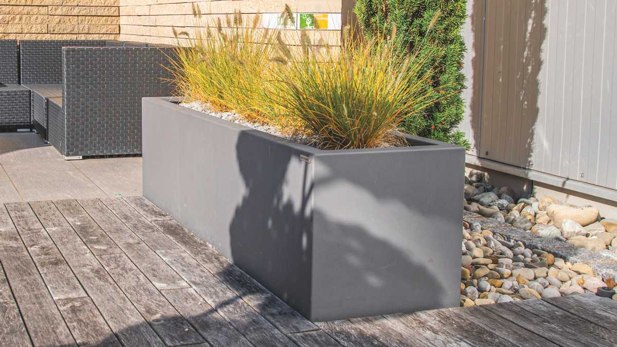 Großer grauer Blumenkasten mit Ziergräsern steht auf Holzterrasse neben schwarzen Flechtmöbeln. Im Hintergrund sind Kieselsteine und eine helle Backsteinmauer zu sehen.
