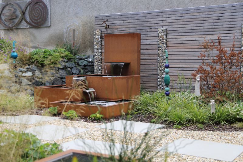 Ein quadratischer Brunnen aus rostig wirkendem Metall lässt Wasser fließen, umgeben von Steinen, Pflanzen und einer Holzwand in einem gepflegten Garten mit dekorativen Skulpturen.