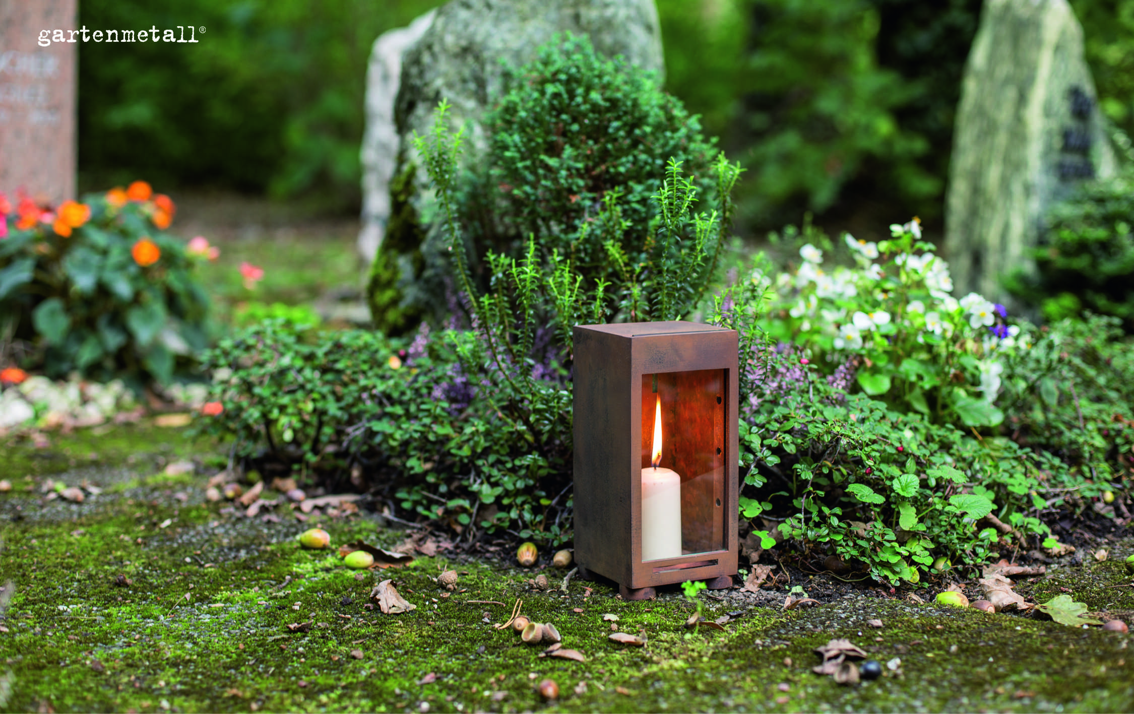 Eine Kerze brennt in einer Metalllaterne, umgeben von grünen Pflanzen und Blumen auf einem moosbewachsenen Boden, neben Grabsteinen. Text: 'gartenmetall®'.