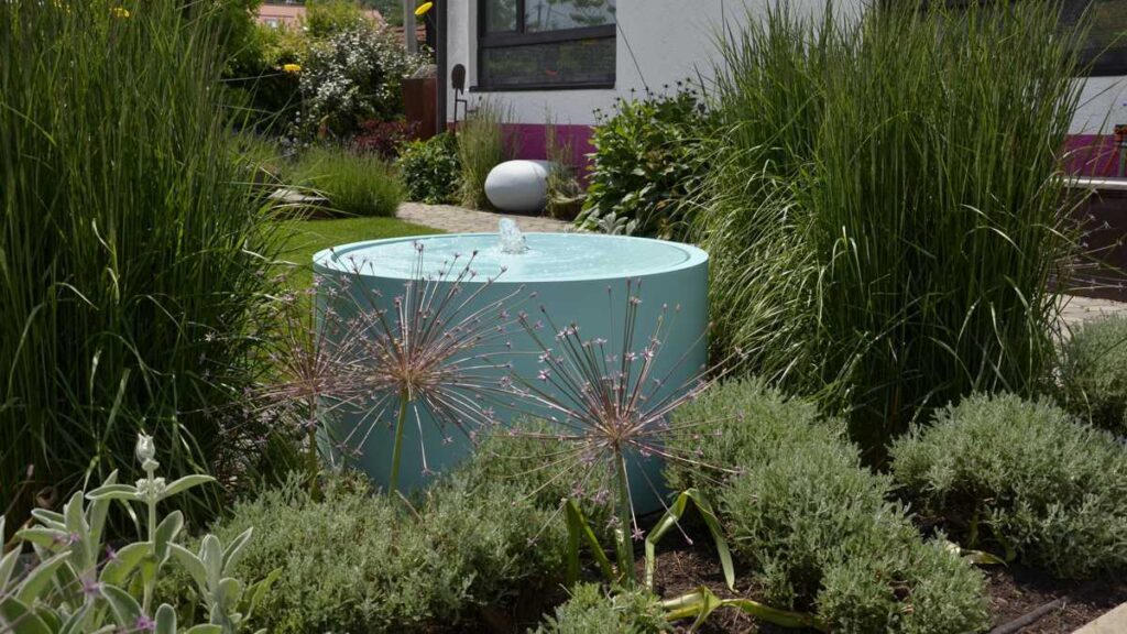 A round, turquoise fountain gently bubbles in the middle of a garden, surrounded by tall grasses and purple-flowering plants near a modern residential building.
