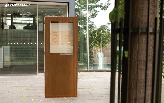Holzkasten steht auf Steinboden, zeigt Dokumente hinter Glas, im Hintergrund moderne Glaswand eines Gebäudes. Text am Eingang: 'gartenmetall®'.