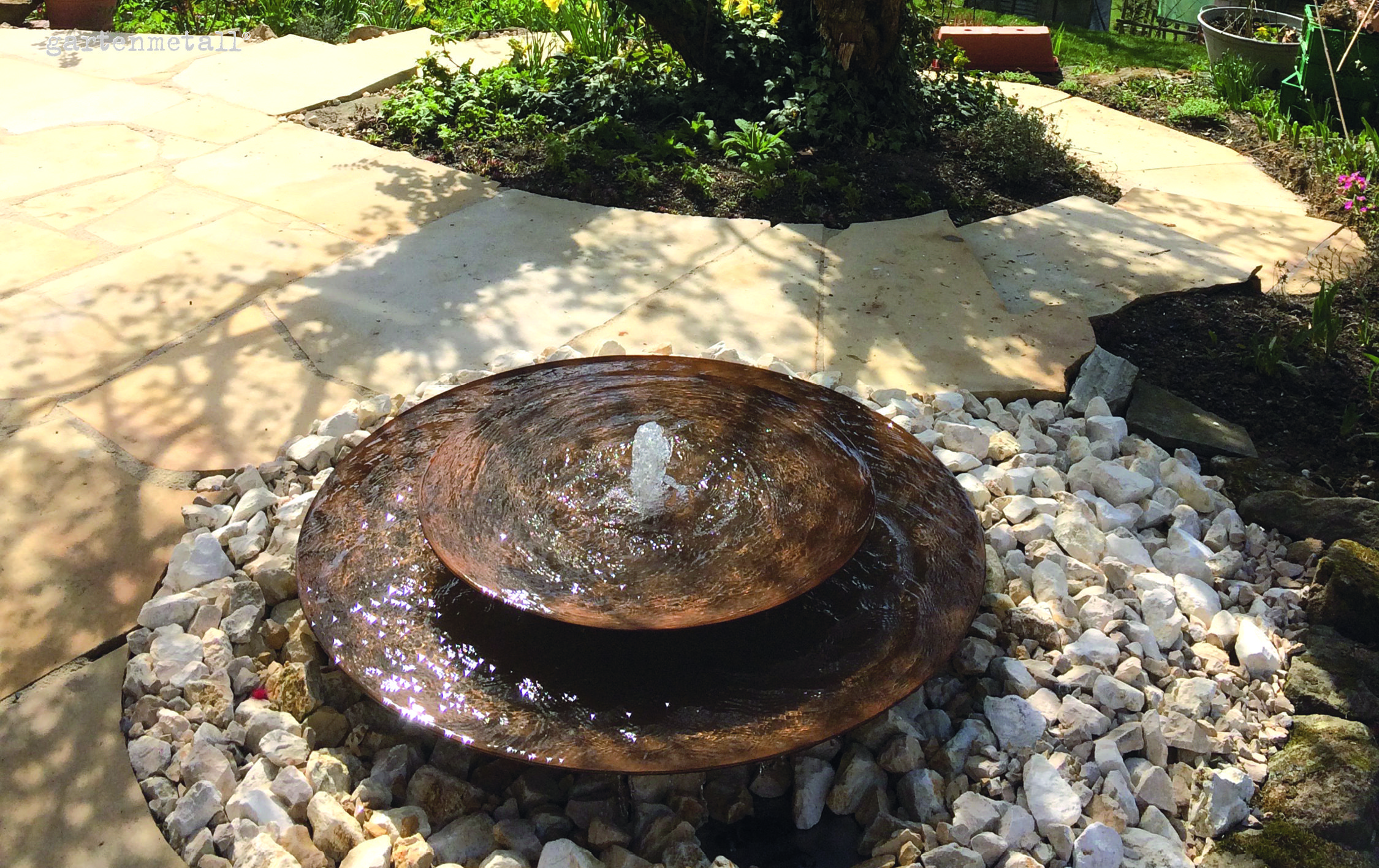 Ein metallener Gartenbrunnen sprudelt Wasser in einem Kreis aus weißen Steinen. Er steht auf einer Pflastersteinterrasse, umgeben von üppigem Grün und blühenden Pflanzen. Text: 'gartenmetall'.
