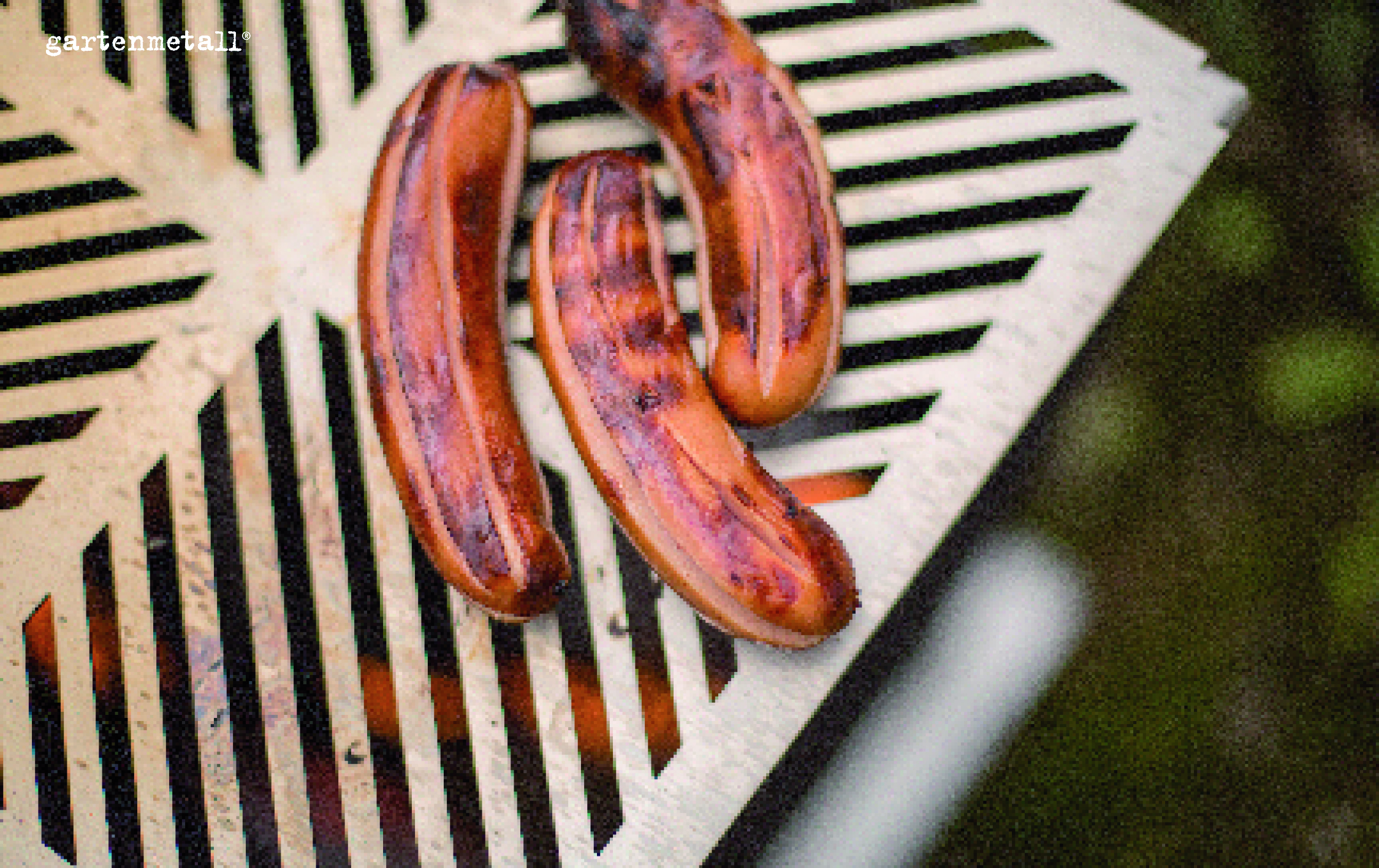 Grillwürste brutzeln auf einem gestreiften Metallrost. Im Hintergrund unscharf erkennt man Vegetation. Der Text 'gartenmetall©' ist oben links sichtbar.