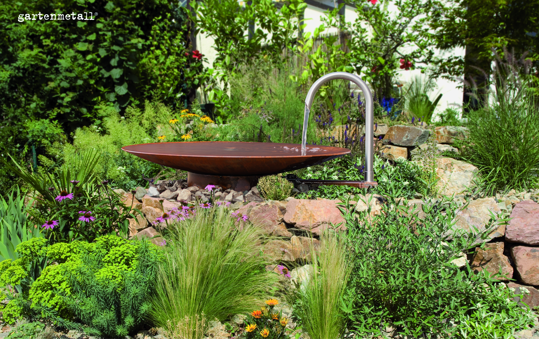 Eine metallene Wasserschale wird über einen gebogenen Wasserhahn befüllt. Sie steht inmitten üppiger Gartenvegetation mit vielen Blumen und ist von Natursteinen umgeben. Text: „gartenmetall®“.