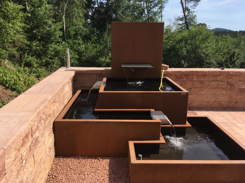 Ein mehrstufiger, rechteckiger Brunnen aus rostfarbenem Metall, fließendes Wasser, eingebettet in eine Steinmauer, mit umgebender grüner Vegetation und rotem Kiesboden.