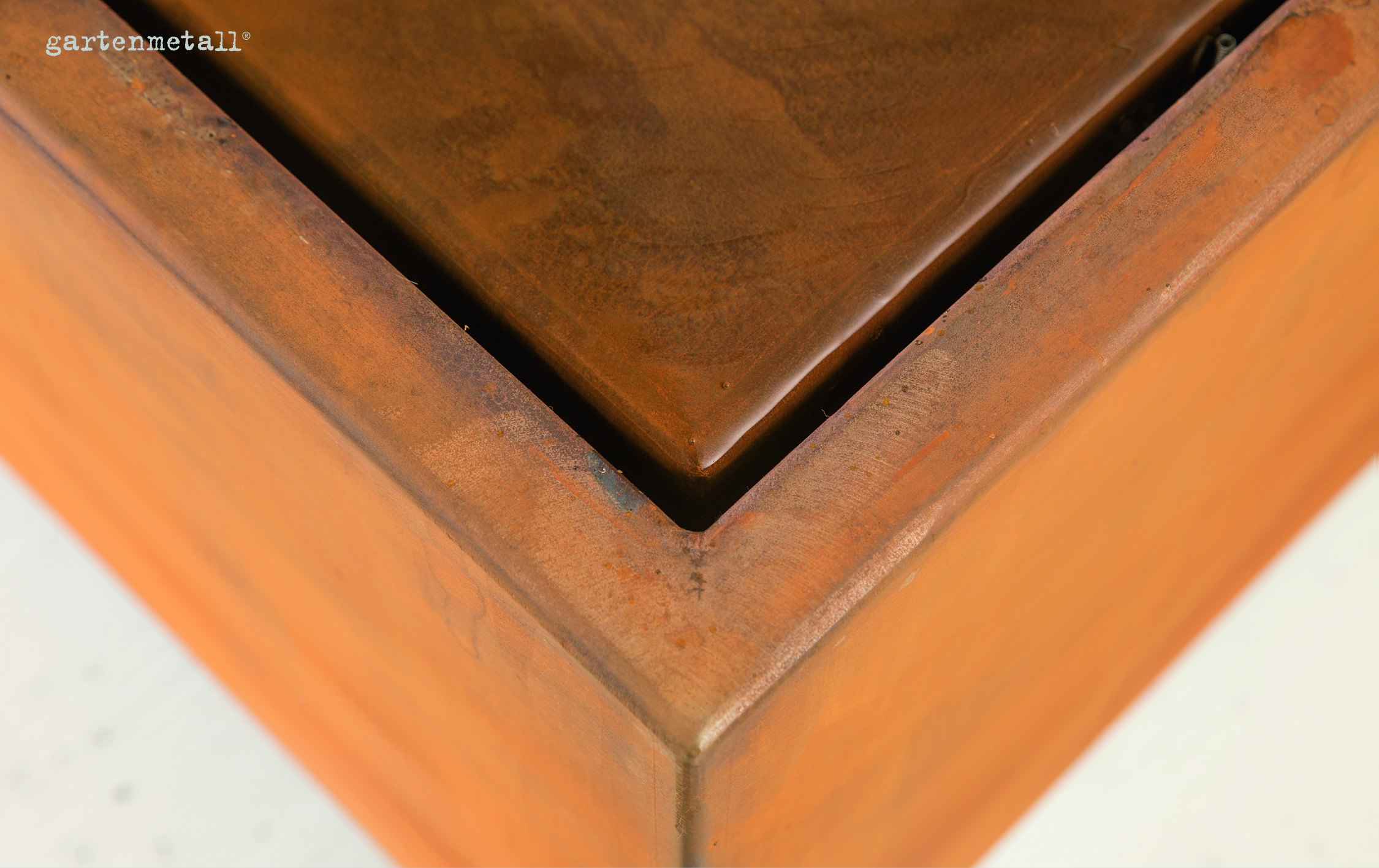 Metallelement bildet eine Ecke, erscheint rostig. Kein sichtbares Geschehen. Befindet sich in einem hellen, wahrscheinlich industriellen Umfeld. Text lautet: 'gartenmetall®'.