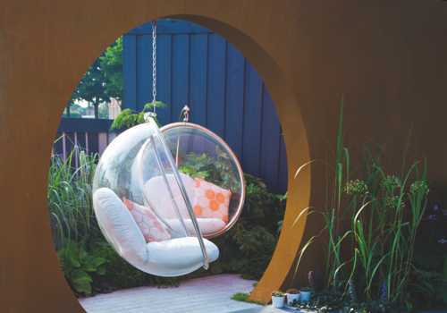 Eine transparente Hängesessel schwingt in einem kreisförmigen Rahmen, umgeben von grünen Pflanzen und Kissen. Im Hintergrund ist ein blauer Holzzaun zu sehen.