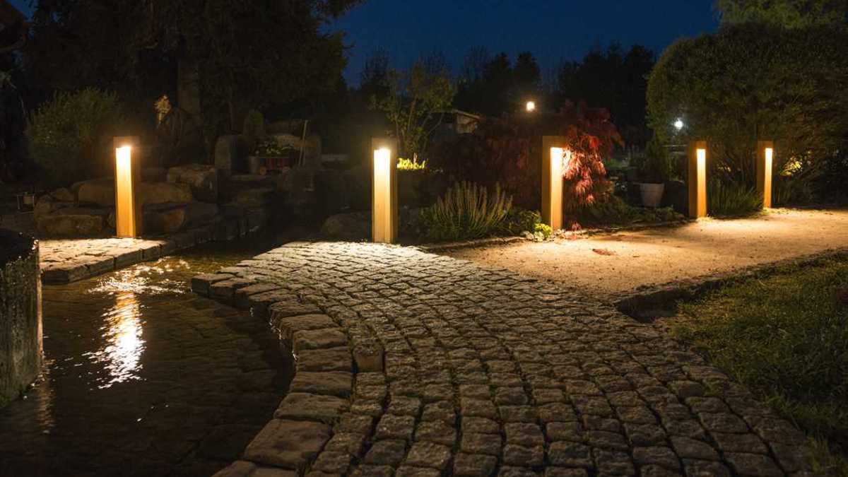 Leuchtpfähle strahlen warmes Licht ab, beleuchten einen geschwungenen gepflasterten Weg neben einem kleinen Bach in einem nächtlichen Garten mit dichten Büschen und Bäumen im Hintergrund.