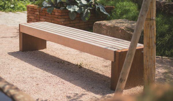 Eine Holzbank steht auf einem Kiesweg, umgeben von Grünpflanzen und Ziegelmauern. Im Hintergrund sind Schattenbereiche und ein sonniger Garten erkennbar.