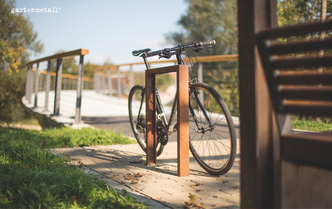 Fahrrad steht an einem Metallständer auf einem gepflasterten Weg, im Hintergrund eine Brücke aus Holz und Metall, umgeben von Bäumen. Text: 'gartenmetall'.