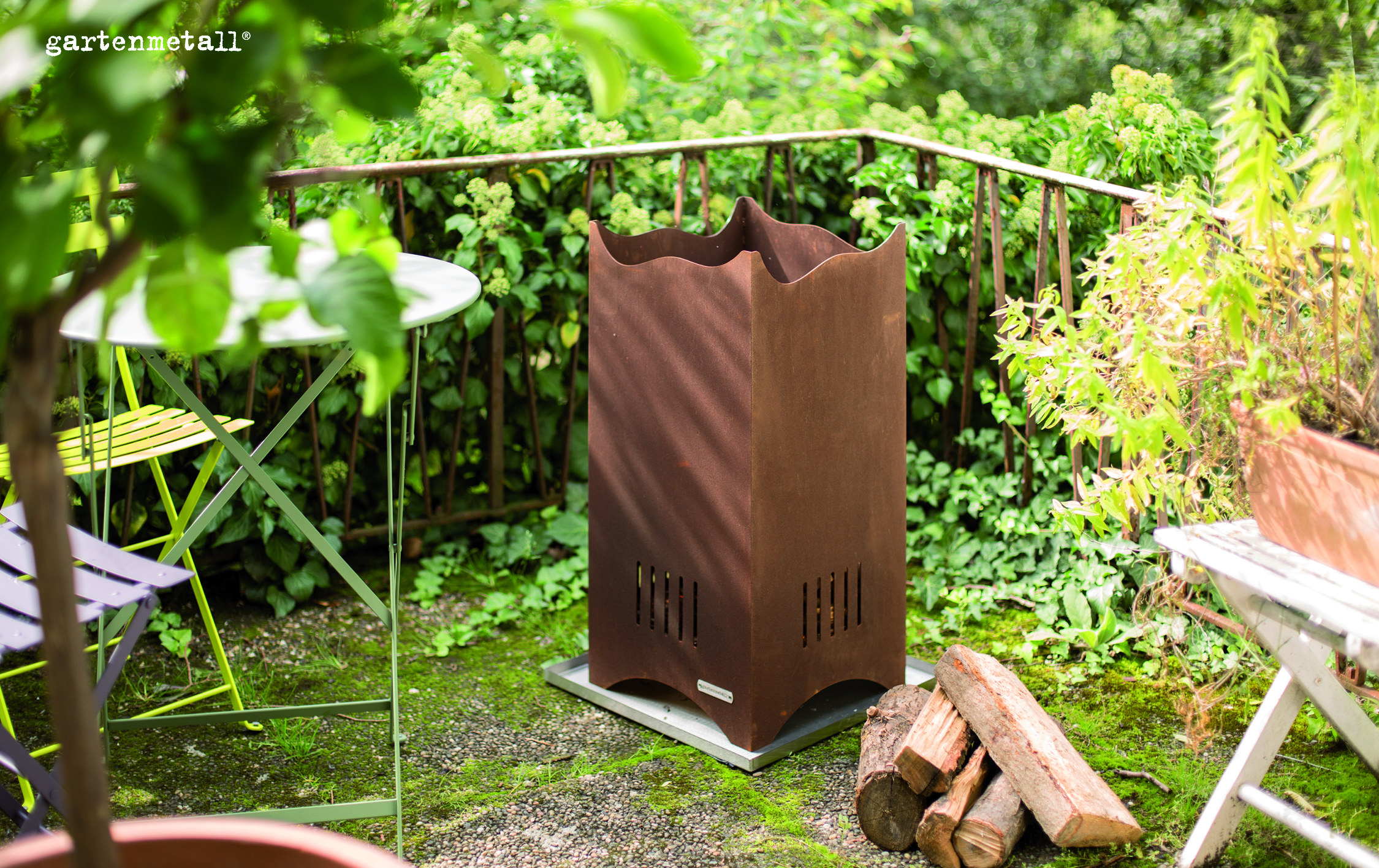 Ein rostfarbener Metallofen steht auf einer Terrasse, umgeben von grünen Pflanzen. Ein Stapel Holzscheite liegt daneben. Text: 'gartenmetall®'.