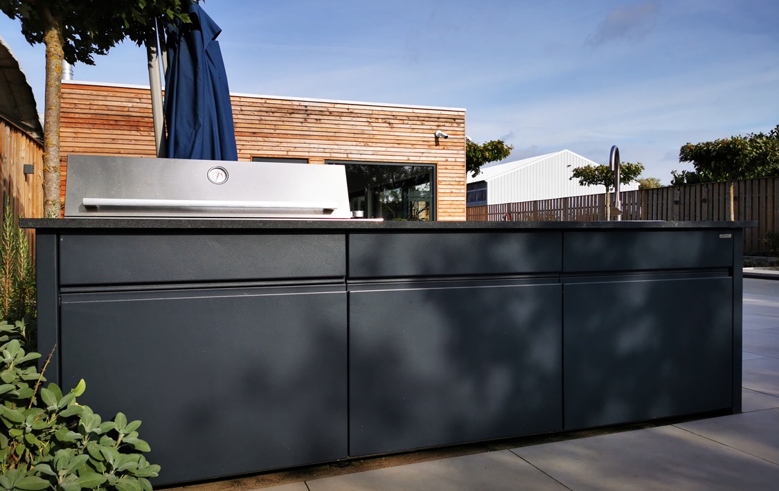 Ein großer, dunkelgrauer Außenküchenschrank steht auf einer Terrasse vor einem modernen Gebäude mit Holzverkleidung, daneben ein Grill und Pflanzen. Ein Sonnenschirm ist im Hintergrund zu sehen.