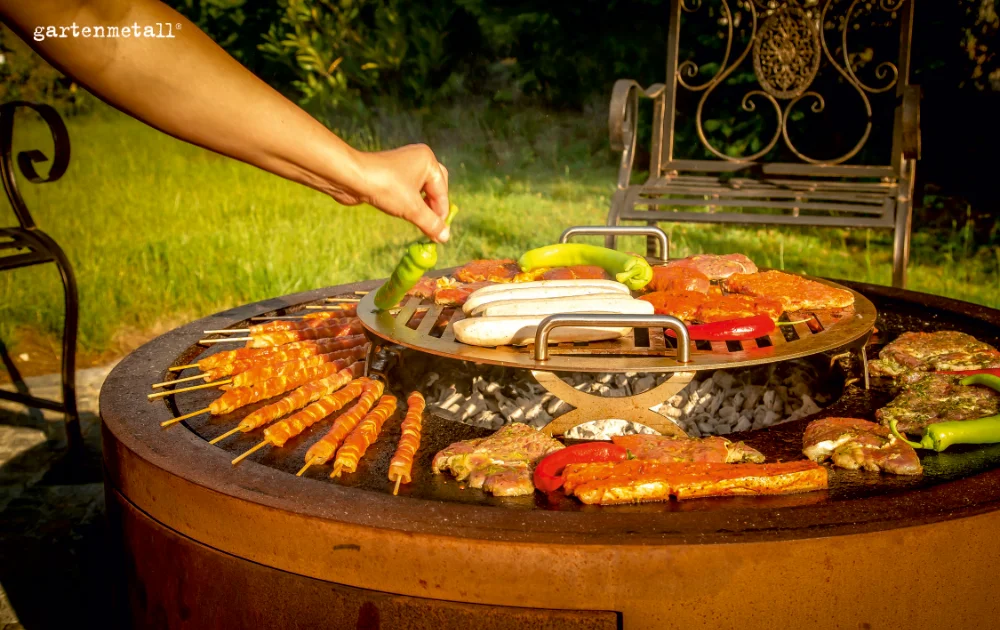 Cortenstahl-Feuerstelle TROJA mit Grillplatte, Grillrost, Fleischspießen, Würstchen und Gemüse im Garten