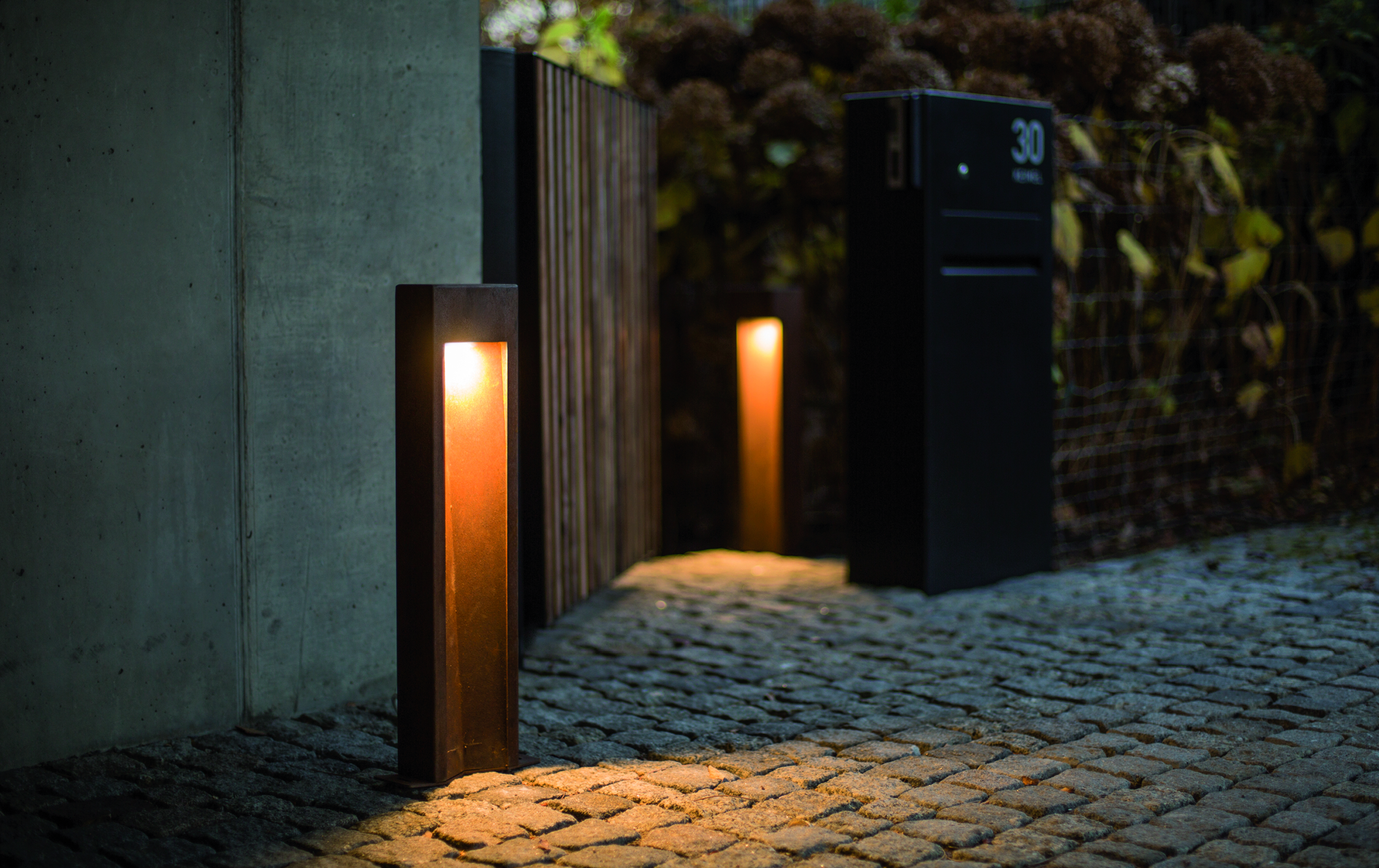Leuchte erhellt den gepflasterten Weg in einem abendlichen Garten, neben einem schwarzen Briefkasten mit der Aufschrift „30“ auf einer Mauer mit Holzzaun und Pflanzen im Hintergrund.