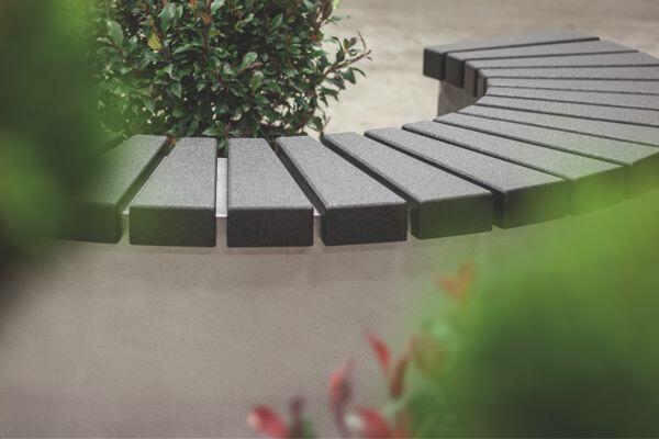 A curved, grey bench stands in a garden area, surrounded by green shrubs and plants, on a paved ground.