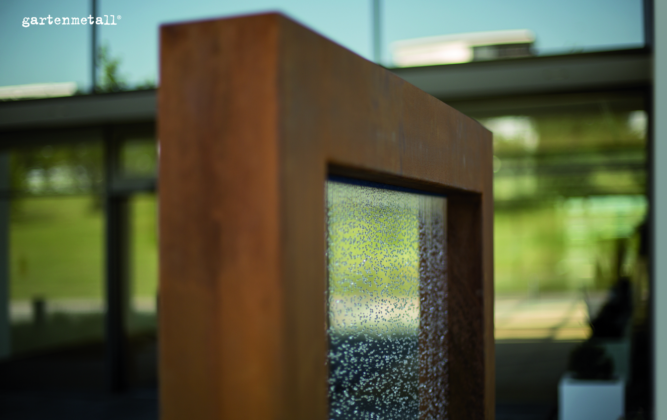 Eine rostbraune Metallskulptur mit eingelassener, mit Wassertropfen bedeckter Glasfläche steht in einem modernen, verglasten Gebäude. Oben links steht der Text: „gartenmetall®“.