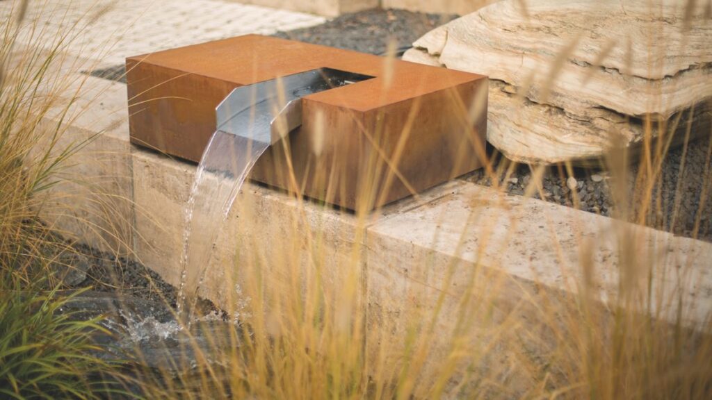 Ein rechteckiger Metallbrunnen mit Wasserstrahl in einer natürlichen Umgebung, umgeben von hohem Gras und Felsen, fließt über einen Betonrand in einen flachen Wasserlauf.